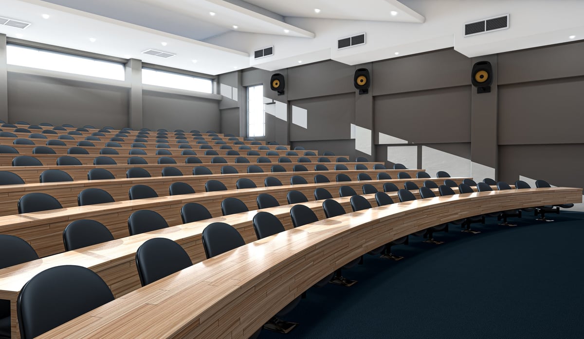 An interior of an empty lecture hall auditorium with rows of curved wooden desks and chairs lit by morning sunlight
