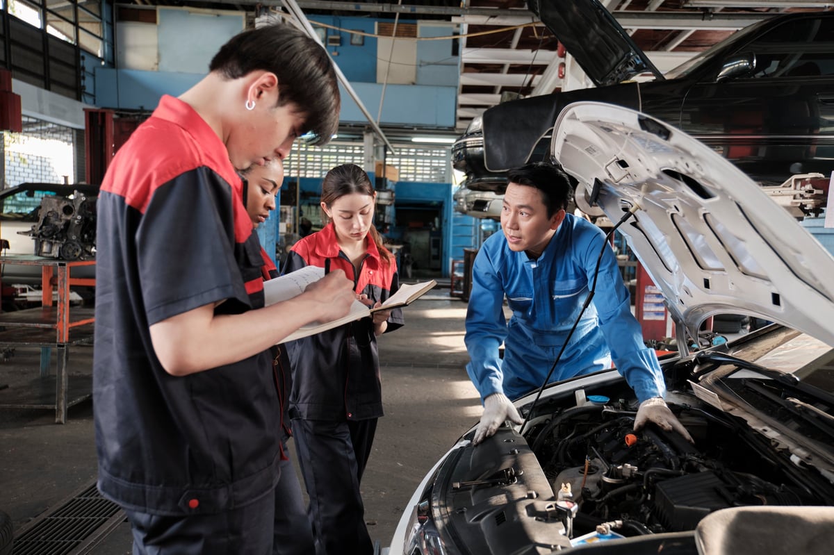 Asian male professional automotive engineer supervisor describes car engine maintenance and repair work with mechanic worker staffs team